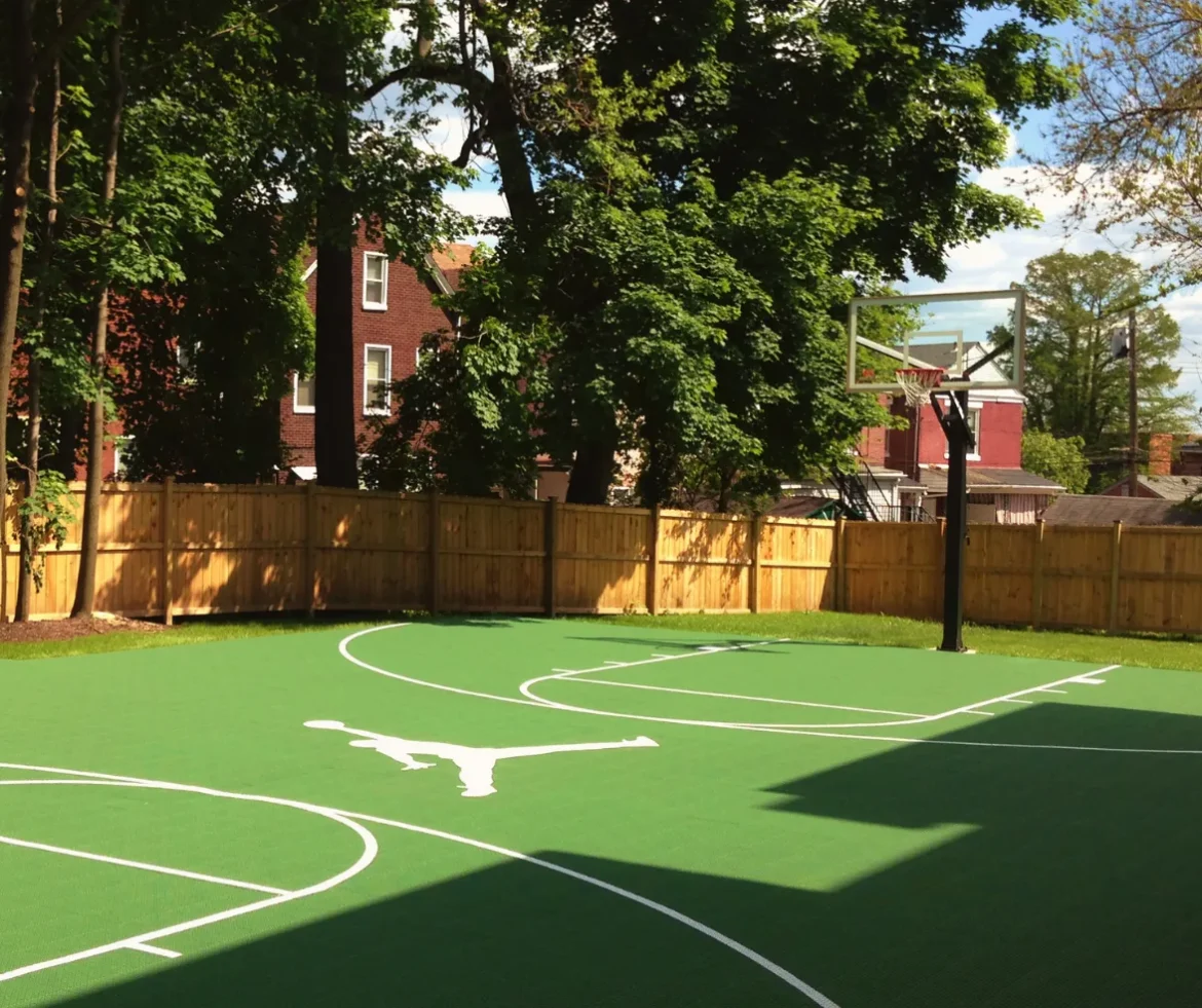 Backyard basketball court with green FlexCourt modular tiles, white game lines, and hoop in a fenced residential yard.