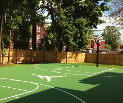 Backyard basketball court with green FlexCourt modular tiles, white game lines, and hoop in a fenced residential yard.